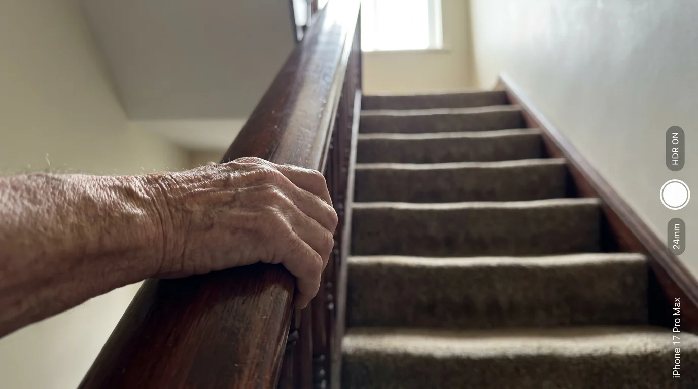 A close-up photo of a person's hand tightly gripping a wooden stair railing, with a steep flight of stairs in the blurred background.