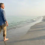 A retiree walks barefoot along a serene, sun-drenched beach with emerald water and white sand under a clear morning sky.