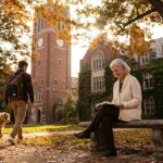 A senior woman sitting on a university bench smiling at a student, with historic brick campus buildings and autumn trees in the background.