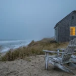 A weathered coastal cottage on a grassy dune at dusk, showing peeling paint on wooden chairs and a view of the gray ocean.