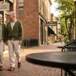 An active senior couple walks down a beautiful, tree-lined historic street with brick buildings and a bakery in the background.