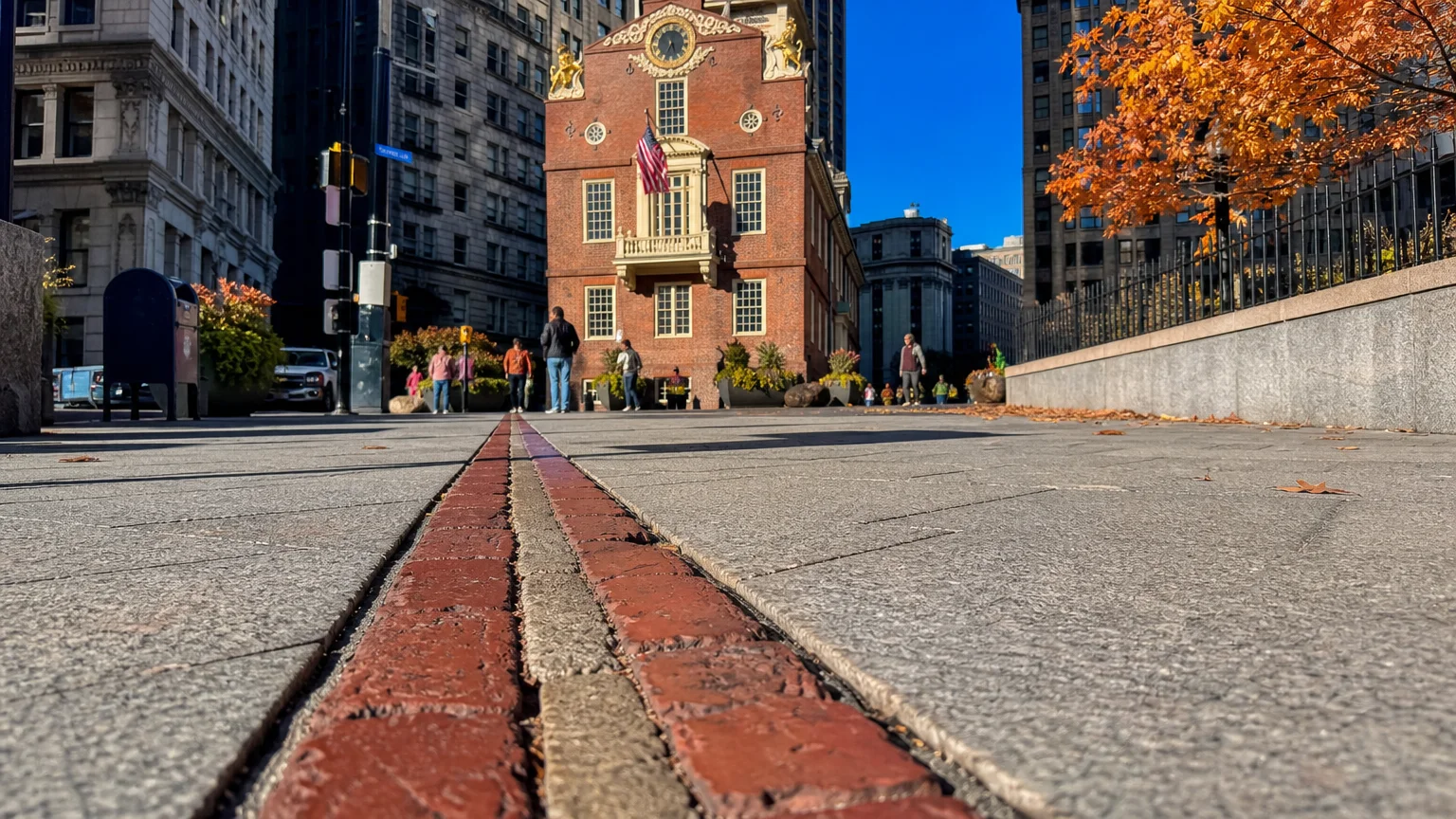 A close-up of the red brick Freedom Trail line on a Boston sidewalk leading toward the historic Old State House.