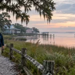 A woman walking along a path by a coastal marsh and houses at dawn with a shrimp boat on the water.