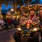 Older couple wearing Santa hats waves from a golf cart decorated with Christmas lights during an evening holiday parade.