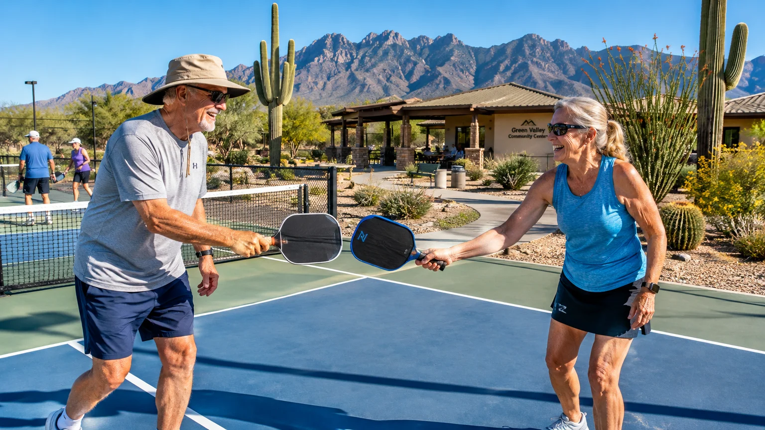 Senior man and woman tapping pickleball paddles on an outdoor court with desert mountains and saguaro cacti behind.